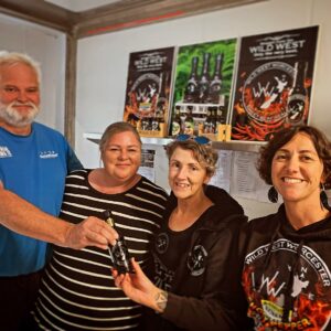 A group of four people standing in a row, who are smiling. On the far left is a tall man with white hair and a beard, and he is being handed a bottle of sauce by a smiling woman on the far right. There are posters saying 'Wild West Worcester' in the background.